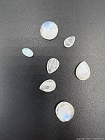 Seven polished moonstones displayed on a black surface showing varying shapes and a blue sheen, typical of moonstone.