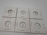 Six small loose opal gemstones secured in individual cardboard holders with clear plastic windows for display and protection.