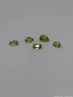Five light green faceted peridot gemstones arranged on a grey background, showing variety of cuts including oval and pear shapes.