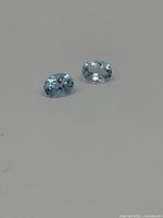 Two oval light blue faceted gemstones on a white background, showing shape, size and color