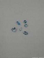 Six loose topaz gemstones displayed on a plain surface, showing various cuts and light blue tones.