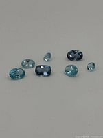 Seven loose topaz gemstones arranged on a flat surface showing different shapes and shades of blue.