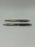 Two ballpoint pens laid flat on a white background, showing the full length and design details of a matte silver vintage-style pen and a textured Cross pen.