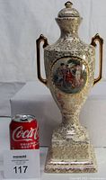 Front view showing entire urn with gold-leafed handles and lid, ivory body, and painted Victorian scene on front.