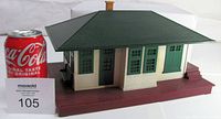 View of Lionel No. 132 model train station, showing the front and side exterior with cream siding, green framed windows and doors, green roof, and maroon base with platform steps. Coca-Cola can for scale, measuring approximately 12" x 8.25" x 6.5".