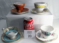 Four teacups and matching saucers arranged around a Coke can for scale, showing varied floral and gold patterns.