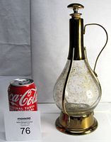 Side view showing overall size of the decanter compared to a soda can. Bottle has bulbous glass body with gold decorating, brass neck and base with handle and stopper.