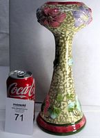 Full view of tall hourglass-shaped ceramic vase beside a soda can for scale. Mottled beige background with raised floral decoration in pink, purple, and green. Glossy glaze.