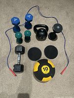 Top-down photo showing two blue 8 lb dumbbells, two green 5 lb dumbbells, black kettlebell, black 10 lb dumbbell, black 15 lb dumbbell, two black sliders, yellow and black 10 lb medicine ball, and blue jump rope.