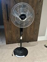 Front view of black oscillating pedestal fan with metal blade cage, control panel, and adjustable stand on a carpeted floor.