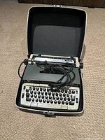 Electric typewriter in open hard carrying case with power cord over keyboard, placed on carpeted floor