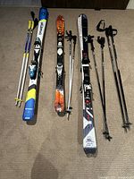 Three pairs of skis with bindings and multiple pairs of ski poles laid out on a carpet showing brands and condition