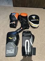 Boxing gloves and two punch catchers arranged on floor; Everlast grey and black gloves and Kimurrawear black and orange gloves visible along with punch catchers.