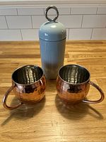 Light blue cocktail shaker standing upright between two copper Moscow Mule mugs on a wooden surface with white tile backsplash.