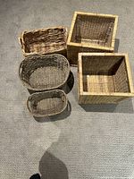 Five wicker baskets on a carpeted floor showing varied sizes and shapes, including square and oval natural wicker baskets.