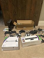 Xbox 360 console standing upright with two wireless controllers and manuals on floor; boxed Kinect sensor and floorstand next to the console; various cables and power supply laid out