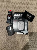 Top-down view of all items laid out: chrome metal hand warmer, original packaging box, lighter fluid bottle, black fabric pouch, white accessory tool.