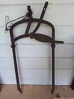 Antique cast iron hay bale hook barn implement with rusted patina leaning against white wall.
