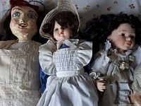 Three dolls including one with painted face and red hair net, one porcelain doll with brown wig and blue dress with white apron, and one porcelain doll with curly black hair in white floral dress.