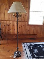 Full height view of the metal floor lamp with beige fabric lamp shade, standing against a wooden wall on a dark rug.