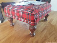 Close-up of the rectangular padded foot stool with red tartan upholstery and turned wooden legs on a hardwood floor