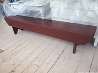 Photo of a long vintage rectangular wooden bench painted red, placed on a floor with visible wear around the bench surface and legs.