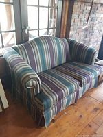 Photo of a modern striped pattern loveseat with rolled arms and skirted base, placed near a window and wooden table.