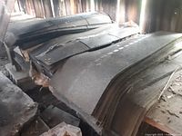 Photo showing multiple large black asphalt roofing shingles stacked in piles inside a storage area with wooden walls.
