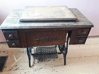 Full view of wooden sewing machine cabinet with veneer top showing wear, drawers, and cast iron legs.
