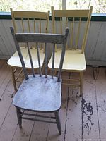 Photo shows three vintage wooden chairs: two painted yellow and one painted white with visible wear and distress. Chairs have spindle backs and wooden seats, placed on a rustic floor.