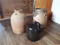 Three vintage crocks including a small brown jug with handle, a medium beige crock with embossed handles, and a larger vase-shaped crock in beige. Displayed on wooden floor.