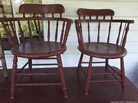 Pair of vintage red painted captain's chairs showing round seats, arched spindle backs and armrests, with visible paint wear and a crack in one seat.
