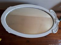 Oval white-framed mirror with beveled glass and Fleur de Lis carved details resting on a wooden floor.