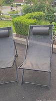 Two black mesh canva beach deck chair recliners with attached headrest cushions, upright position, outside on pavement.