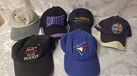 Six baseball caps arranged on table showing various sports and brand logos