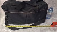 Black duffel bag measured next to a water bottle and tape measure, showing approximate size 19x10x10 inches