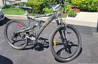 Photo of a gray Schwinn Vertex full suspension mountain bike parked on asphalt with greenery in background. Condition shows light use, with some wear visible on the rear tire.