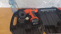 Image shows an orange and black Black and Decker cordless drill inside a black plastic carrying case, alongside a plastic water bottle for scale. The case has compartments with some screws present.