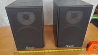 Pair of Braxton bookshelf speakers placed side by side showcasing front fabric covers with tweeter and woofer visible beneath. A measuring tape laid horizontally in front shows size details.