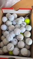 Box containing about 50 mixed brand golf balls including Titleist, Callaway and others, mostly white, some yellow.