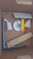 Box containing various miscellaneous hardware and household items including lamp socket, scraper, paintbrush, sanding tool, and corner braces.