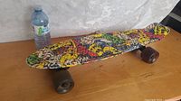 Side angled view of a used Penny skateboard showing colorful graphic deck and four dark wheels on a wooden table with a water bottle in the background.