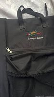 Close-up of black fabric with embroidered Lounge Lizard logo and front zippered pocket of stadium seat.