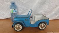 Side view of blue and white 1960s Tonka Jeep toy with water bottle for scale, showing metal body, white plastic seats, whitewall tires, and mild rust.