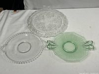 Three glass dishes displayed on a white surface: a large clear platter with floral edge pattern, a clear plate with beaded rim, and a green glass plate with handles.