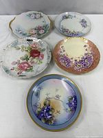Five floral decorative plates arranged on beige surface showing varied floral designs and gold trim.