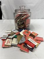 Glass jar filled with vintage matchbooks with some matchbooks spread around it showing variety.