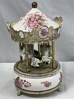 Porcelain and metal merry go round music box decorated with cherry blossom flowers and featuring three carousel horses. Gold-tone metal detailing and pink floral motifs on canopy and base. Close up view angle.