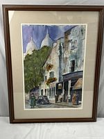 Framed watercolor painting showing European street with buildings, trees, people, and vintage car, signed by artist.