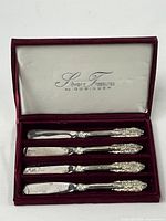 Set of four spreadable cheese knives displayed in maroon velvet-lined box labeled 'Silver Treasures by Godinger'. Each knife has an ornate floral and scroll design on handle.